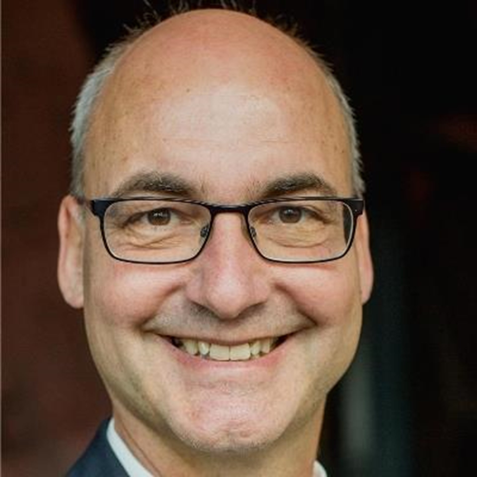 Lächelnder Mann mit grauen Haaren, Halbglatze und Brille, der ein weißes Hemd und eine graue Anzugjacke trägt